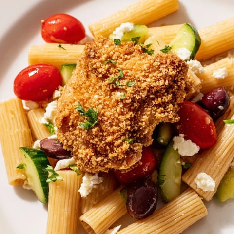 Golden crispy chicken bites rest atop a colorful bowl of pasta tossed with fresh Greek salad vegetables and crumbled feta cheese.