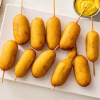 Freshly fried Corn Dogs on a stick, dripping with golden cornmeal batter and served with ketchup and mustard.